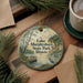 A round ornament with a scenic lake and park landscape, featuring a bird and a dock, is displayed on a wooden surface alongside a cup of coffee and some pine branches.