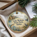 A round ornament with a scenic lake and mountains image, hanging from a string, placed in a box with pine branches.