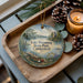 A decorative ornament featuring a scenic lake landscape with a boat, pine cones, and a candle on a wooden tray.