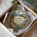 A decorative ornament in a box, featuring a scenic landscape with a lighthouse and a path leading through a forest.