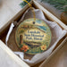 A circular ornament with a landscape scene and text, placed in a box with a white tissue paper lining.