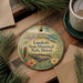 A decorative ornament with a landscape scene and text, placed on a wooden surface next to a cup of coffee and pine branches.