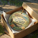 A circular ornament with a landscape scene and text, placed in a cardboard box with a gold string.
