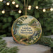 A round ornament with a painting of a scenic landscape, featuring a lake, a bridge, and a bird, hangs in front of a Christmas tree with twinkling lights.