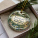 A decorative ornament in a box, featuring a scenic landscape with pine trees and a body of water.