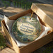 A circular ornament in a brown box, featuring a scenic landscape with pine trees, a lake, and a castle, with the text "Leominster State Forest, Massachusetts" printed on it.