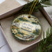 A decorative ornament in a box, featuring a scenic landscape with a lake, a bird, and a house.