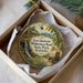 A round ornament with a painting of a wagon traveling down a road surrounded by trees and a waterfall, placed in a box with white tissue paper.