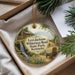 A round ornament with a painting of a cabin and a waterfall, surrounded by greenery, placed in a box with pine branches.