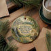 A round ornament with a scenic illustration of a cabin and a waterfall, surrounded by greenery, is displayed on a wooden surface alongside a cup of coffee and some papers.