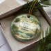 A decorative ornament in a box, featuring a scenic landscape with a lake, trees, and a picnic table.