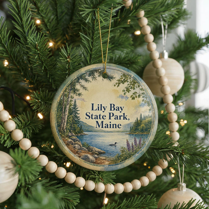 A round ornament hanging on a Christmas tree, featuring a scenic landscape with a lake, pine trees, and a bird in flight.