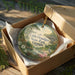 A decorative ornament in a box, featuring a scenic landscape with a deer, flowers, and a bird, along with the text "Little Ocmulgee State Park, Georgia".