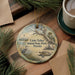 A decorative ornament featuring a serene scene of a bird, a turtle, and a lighthouse, with the text "Little Talbot Island State Park, Florida" displayed on it. The ornament is placed on a wooden surface, surrounded by pine branches and a cup of coffee.