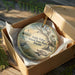 A circular ornament in a wooden box, featuring a scenic landscape with a lighthouse, palm trees, and a bird.