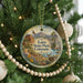 A round ornament hanging on a Christmas tree, featuring a scenic landscape with mountains, a river, and a deer.