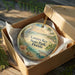 A round ornament in a box, featuring a tropical beach scene with palm trees, a bird, and a turtle.
