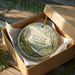 A decorative ornament in a box, featuring a scenic landscape with a river and trees, with the text "Lovers Leap State Park, Connecticut" printed on it.