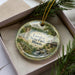 A decorative ornament in a box, featuring a scenic landscape with a river, trees, and a cabin.