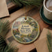 A decorative ornament featuring an illustration of Lowell Heritage State Park in Massachusetts, with a cup of coffee and pine branches in the background.