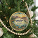 A round ornament hanging on a Christmas tree, featuring a scenic landscape with mountains, a lake, and a boat.