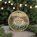 A circular ornament with a scenic landscape of mountains, a lake, and a boat, hanging in front of a Christmas tree with lights.