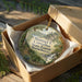 A decorative ornament in a wooden box, featuring a scenic landscape with pine trees, a waterfall, and a mountain range.