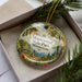 A decorative ornament in a box, featuring a scenic landscape with a mountain, pine trees, and a path leading through the woods.