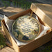 A decorative ornament in a wooden box, featuring a vintage-style illustration of a house surrounded by nature, with the text "Marlorie Kinnan Rawlings Historic State Park, Florida" displayed on it.