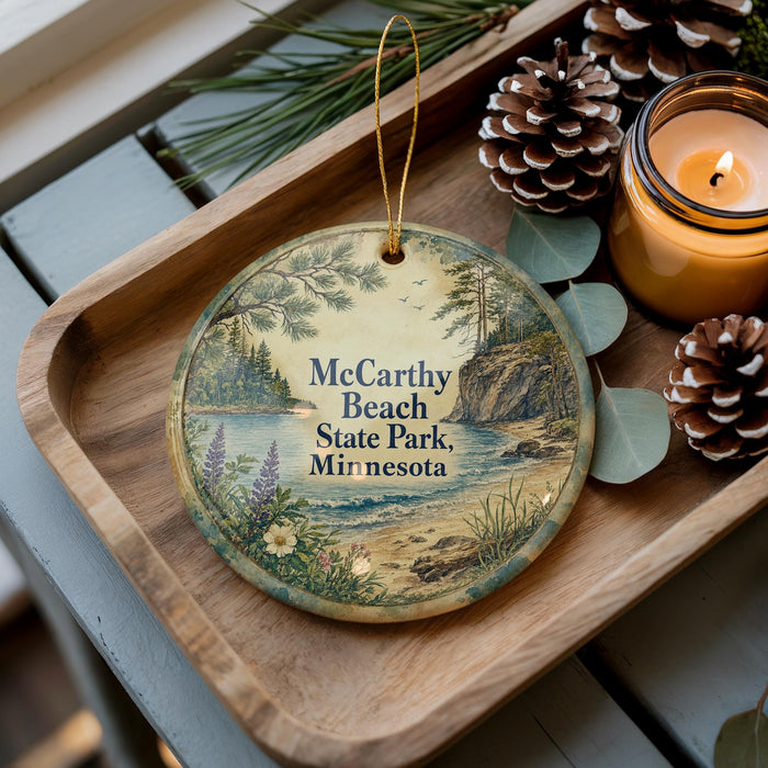 A decorative ornament with a scenic landscape of McCarty Beach State Park in Minnesota, featuring pine cones, a candle, and greenery.