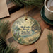 A decorative ornament with a scenic landscape of McCarty Beach State Park, Minnesota, featuring pine trees, a lake, and mountains. The ornament is hanging on a wooden surface, surrounded by pine branches and a cup of coffee.