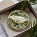 A decorative ornament in a box, featuring a scenic landscape with a waterfall and pine trees.