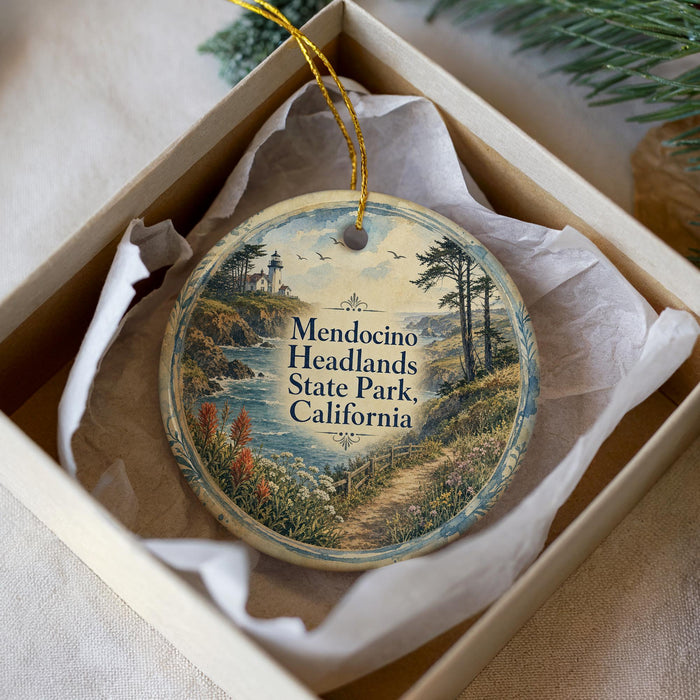 A decorative ornament in a white box, featuring a scenic landscape with a lighthouse, trees, and a path leading to Mendocino Headlands State Park in California.