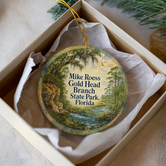 A round ornament with a painting of a scenic landscape, including a river, trees, and a bridge, is displayed in a box.