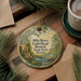 A round ornament with a painting of a forest scene hangs on a wooden surface, accompanied by a cup of coffee and some pine branches.