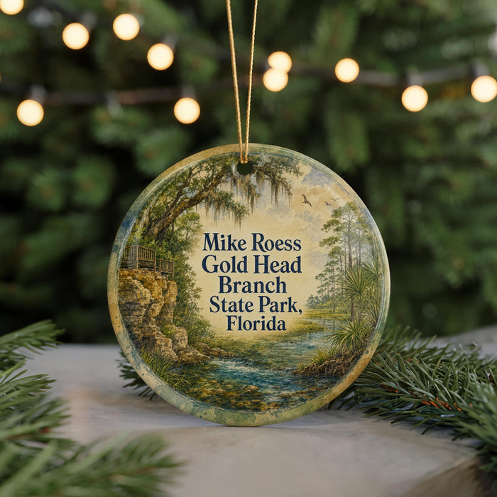 A round ornament with a painting of a scenic landscape, featuring a river, trees, and a bridge, hangs in front of a blurred background with Christmas lights.
