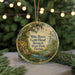 A round ornament with a painting of a scenic landscape, featuring a river, trees, and a bridge, hangs in front of a blurred background with Christmas lights.