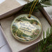A decorative ornament with a scenic landscape of Millers Pond State Park, Connecticut, displayed in a box with pine branches.