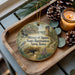 A wooden tray with a decorative ornament featuring a scenic landscape, including a waterfall, a tower, and a path, along with pine cones and a lit candle.