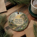 A decorative ornament featuring a scenic landscape with a cabin, trees, and a bridge, hanging on a wooden surface.