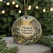 A round ornament with a painting of a scenic landscape, featuring a cabin, trees, and a bridge, hanging in front of a blurred background with Christmas lights.