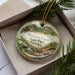 A decorative ornament in a box, featuring a scenic landscape with a river, trees, and a house, along with the text "Mocasin Creek State Park, Georgia".