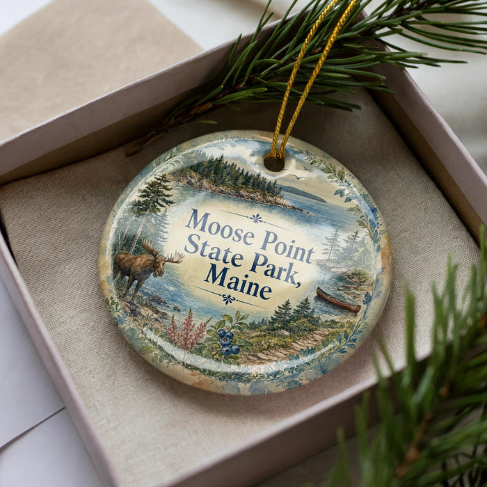 A decorative ornament in a box, featuring a moose and a scenic landscape with pine trees and a body of water.