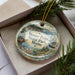 A decorative ornament in a box, featuring a moose and a scenic landscape with pine trees and a body of water.