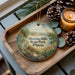 A decorative ornament with a scenic landscape of Moraine Hills State Park, Illinois, featuring a winding path, pine trees, and a lake. The ornament is displayed on a wooden tray, accompanied by pine cones and a lit candle.
