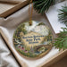 A decorative ornament featuring a scenic landscape with a bridge, water, and flowers, placed in a box with pine branches.