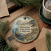 A decorative ornament featuring a scenic landscape of Morro Bay State Park, California, with a bear resting on a rock and a sailboat on the water. The ornament is hanging on a wooden surface, surrounded by pine branches and a cup of coffee.