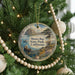 A round ornament hanging on a Christmas tree, featuring a scenic landscape with a mountain, ocean, and a bear.