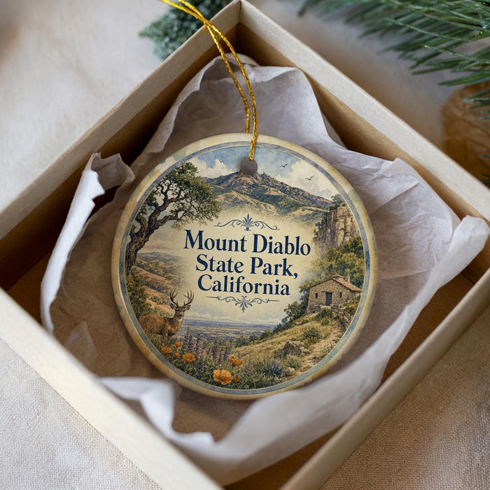 A decorative ornament in a box, featuring a scenic landscape with mountains, trees, and a small building.