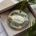 A decorative ornament in a box, featuring a scenic landscape with a castle, trees, and a winding path.